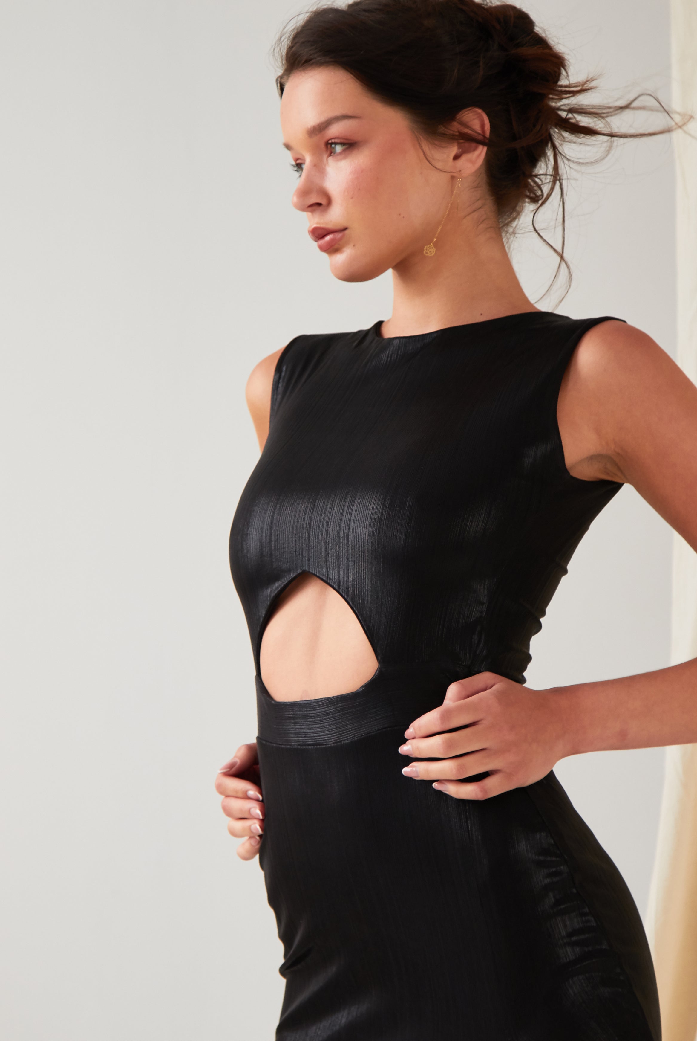 A person wearing the Sarvin Black Cut-Out Maxi Dress with a circular waist cutout stands against a light background, looking to the side with hands on their hips.