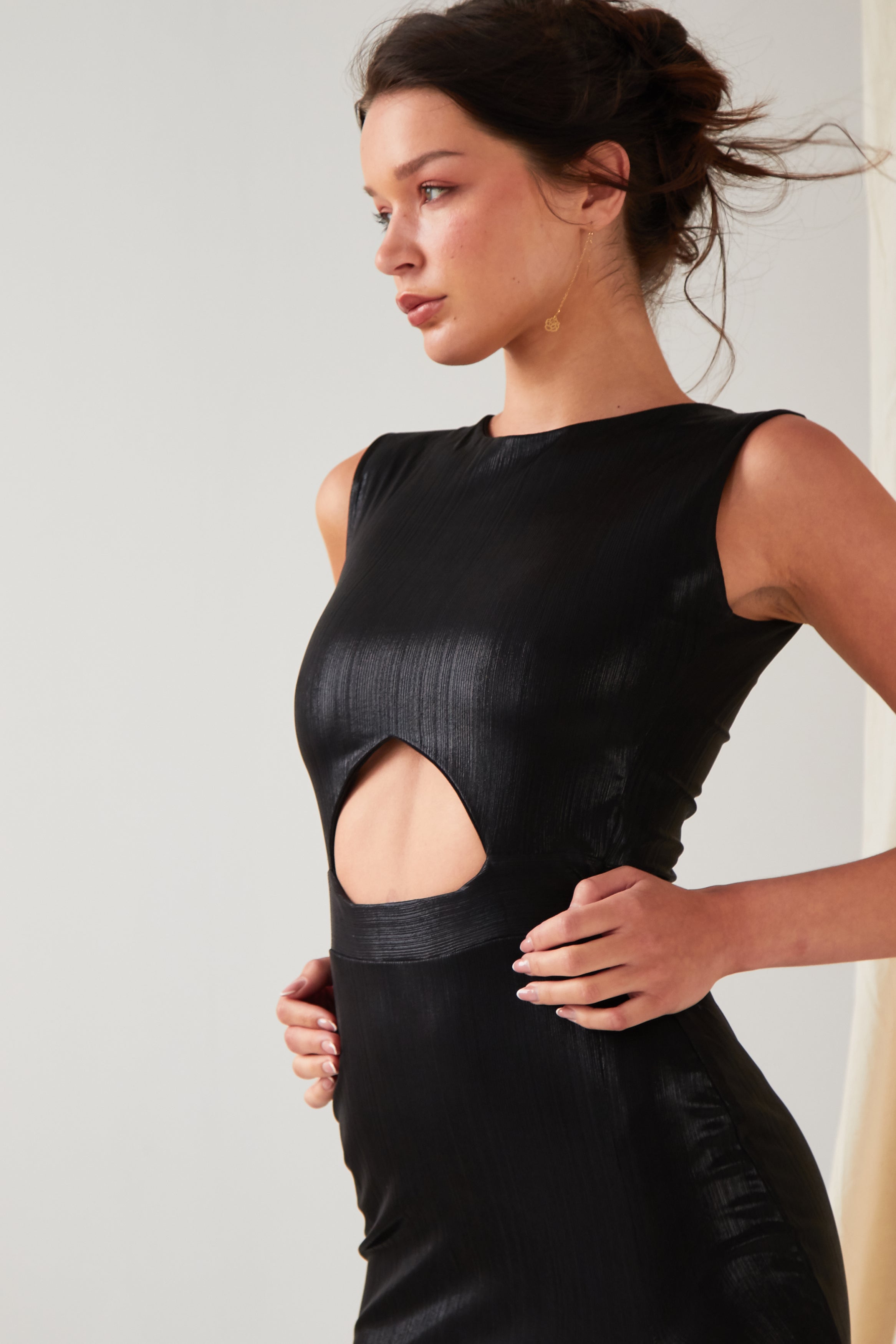 A person wearing the Sarvin Black Cut-Out Maxi Dress with a circular waist cutout stands against a light background, looking to the side with hands on their hips.