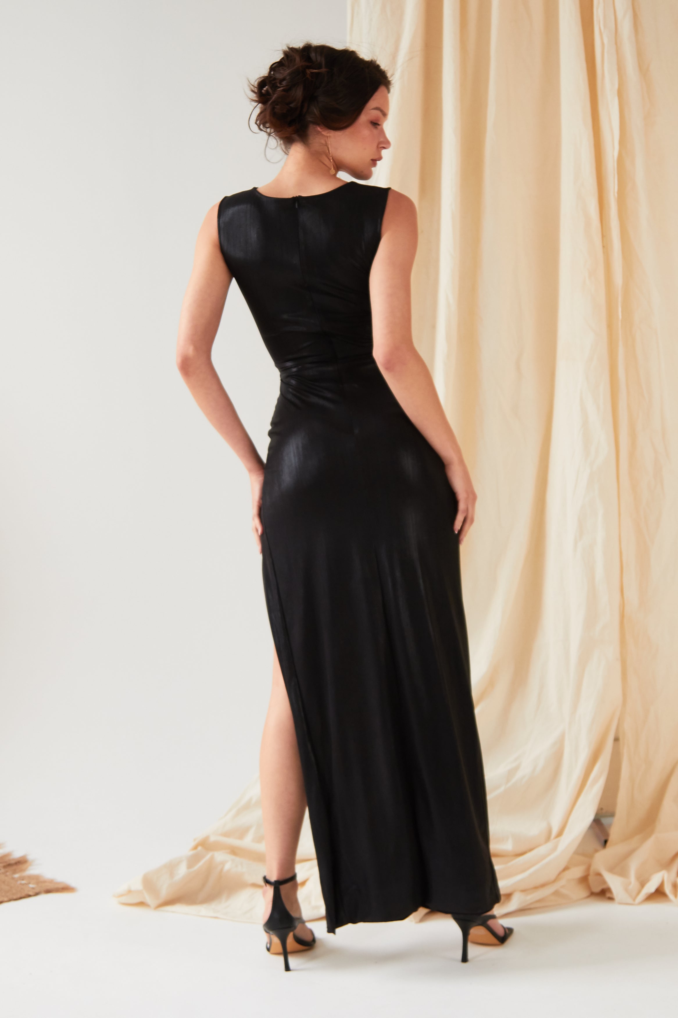 A person models the Sarvin Black Cut-Out Maxi Dress, featuring a side slit and sleek design, standing with their back turned and looking over their shoulder, set against beige draped fabric and a light floor.
