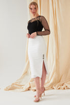 A woman stands before beige draped fabric, wearing a black long-sleeve sheer polka dot top, Sarvin's handmade in the UK White Bodycon Lace Skirt with side buttons and slit, and nude high-heeled sandals.