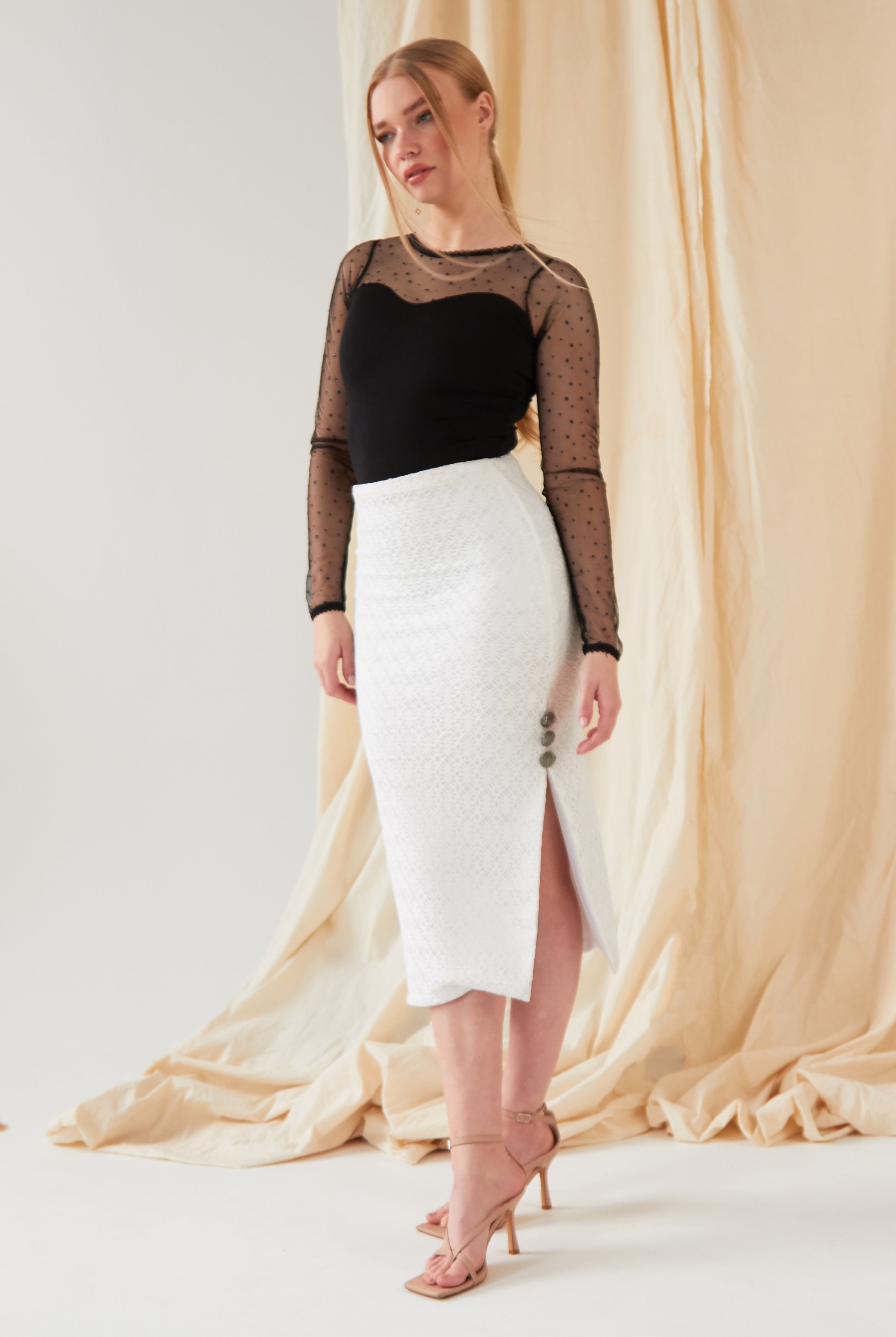 A woman poses before a cream draped backdrop, wearing a black sheer polka dot top and the Sarvin White Bodycon Lace Skirt with side buttons and a slit, finished with nude high-heeled sandals.