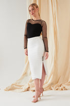 A woman poses before a cream draped backdrop, wearing a black sheer polka dot top and the Sarvin White Bodycon Lace Skirt with side buttons and a slit, finished with nude high-heeled sandals.