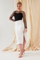 A woman stands indoors in the Sarvin Long-Sleeve Mesh Bodysuit, styled with a white high-waisted skirt featuring a front slit and buttons, nude heels, and a light beige fabric backdrop.