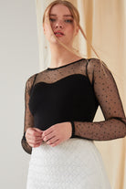 Wearing a black sheer polka-dot top over a black tank, a white lace skirt, and Sarvin Clover Shape 18K Gold Threader Earrings, a person stands before a beige curtain with loose hair framing their face.