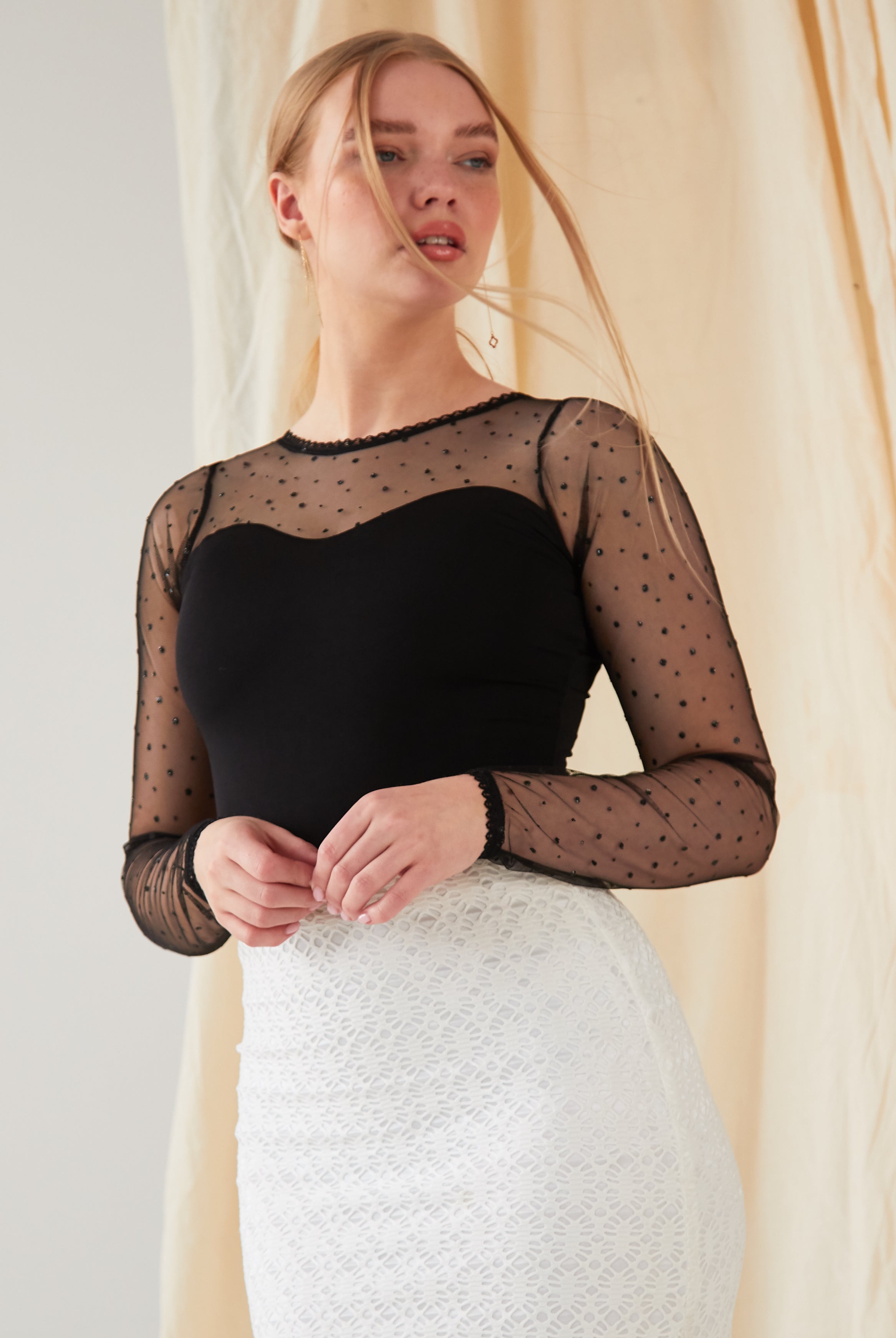 A woman with light hair stands before a beige curtain, wearing the Sarvin Long-Sleeve Mesh Bodysuit with sheer polka dots over a black camisole and a high-waisted white lace skirt, gazing to the side.