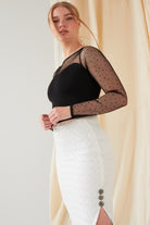 A person stands in profile wearing a black, long-sleeved mesh top with polka dots and the Sarvin White Bodycon Lace Skirt, handmade in the UK, featuring two decorative buttons. Cream-colored fabric hangs in the background.