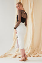A woman poses before a beige fabric backdrop, wearing Sarvin's handmade White Bodycon Lace Skirt (UK) with back slit and buttons, styled with a black sheer polka dot top and nude heels. She gazes over her shoulder.