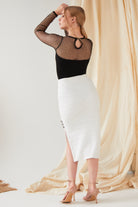 A woman faces away, wearing a black sheer polka dot top and the Sarvin White Bodycon Lace Skirt—handmade in the UK with a high waist, textured fabric, and side slit—paired with nude heels against a cream draped background.