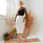 A woman stands indoors on a woven rug, wearing the Sarvin White Bodycon Lace Skirt with a side slit and a black sheer long-sleeve top. Behind her are pampas grass, a dreamcatcher, and potted plants.