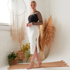 A woman stands indoors on a woven rug, wearing a black sheer long-sleeve top and the Sarvin White Bodycon Lace Skirt—high-waisted, handcrafted in the UK with side buttons and a slit. Pampas grass, dreamcatcher, and plants decorate the scene.
