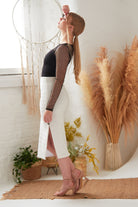A woman in profile wears the Sarvin Long-Sleeve Mesh Bodysuit with a white skirt and nude heels, posing with one hand on her forehead. Pampas grass and a dreamcatcher are visible in the background.