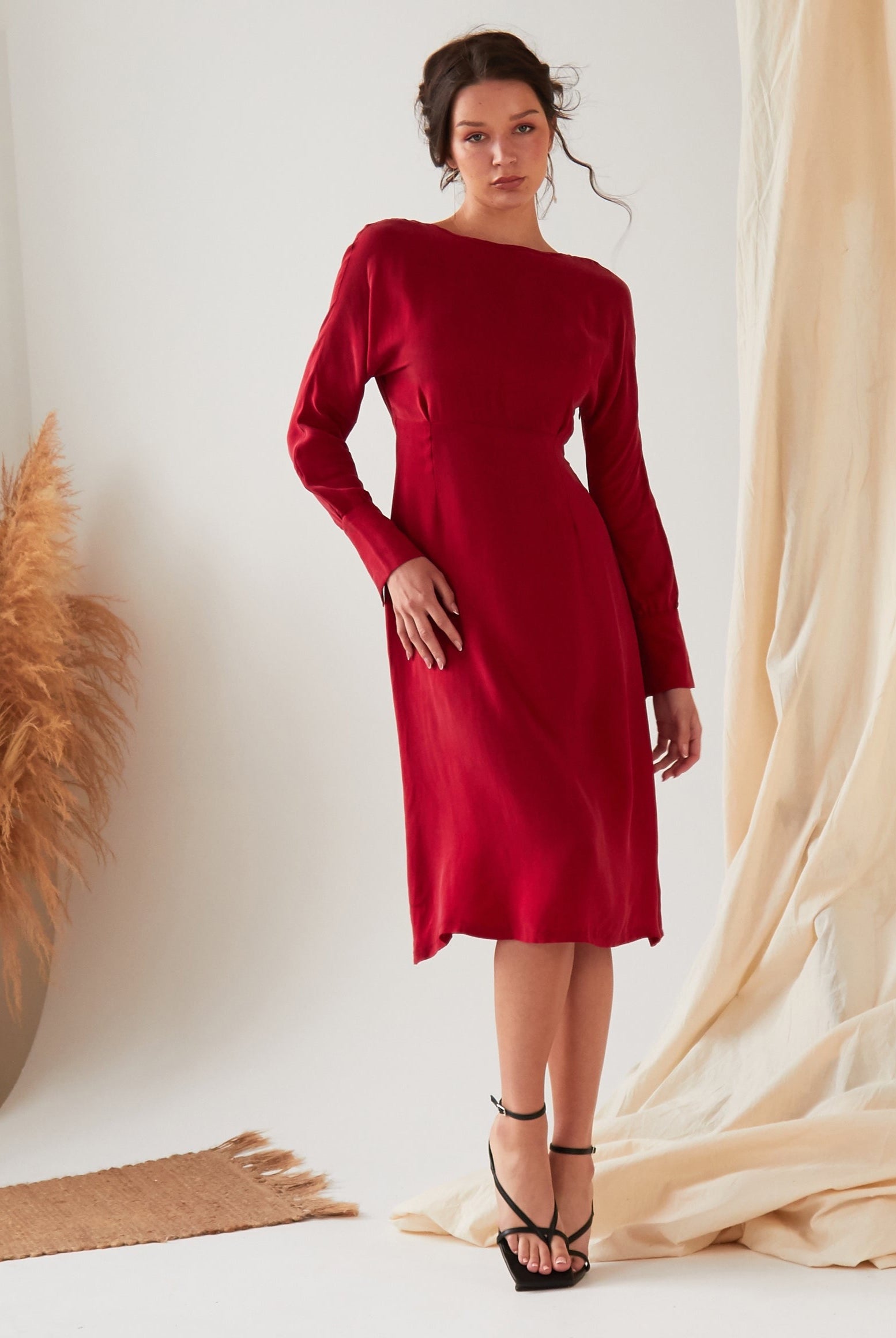 A woman wears the Sarvin Long Sleeve Red Midi Dress with black strappy heels, posing indoors against a neutral backdrop with beige curtains and dried pampas grass.