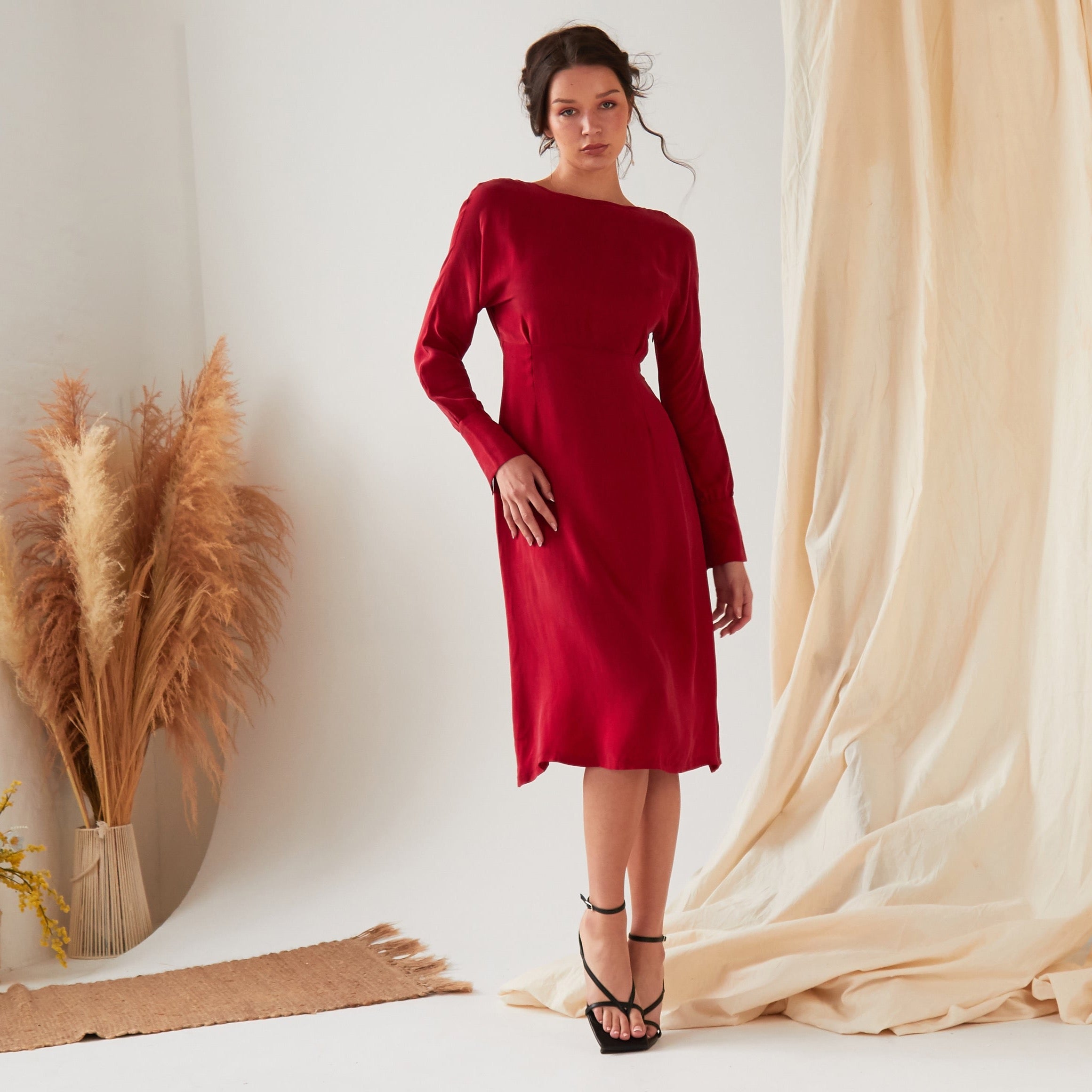 A woman wears the Sarvin Long Sleeve Red Midi Dress with black strappy heels, posing indoors against a neutral backdrop with beige curtains and dried pampas grass.