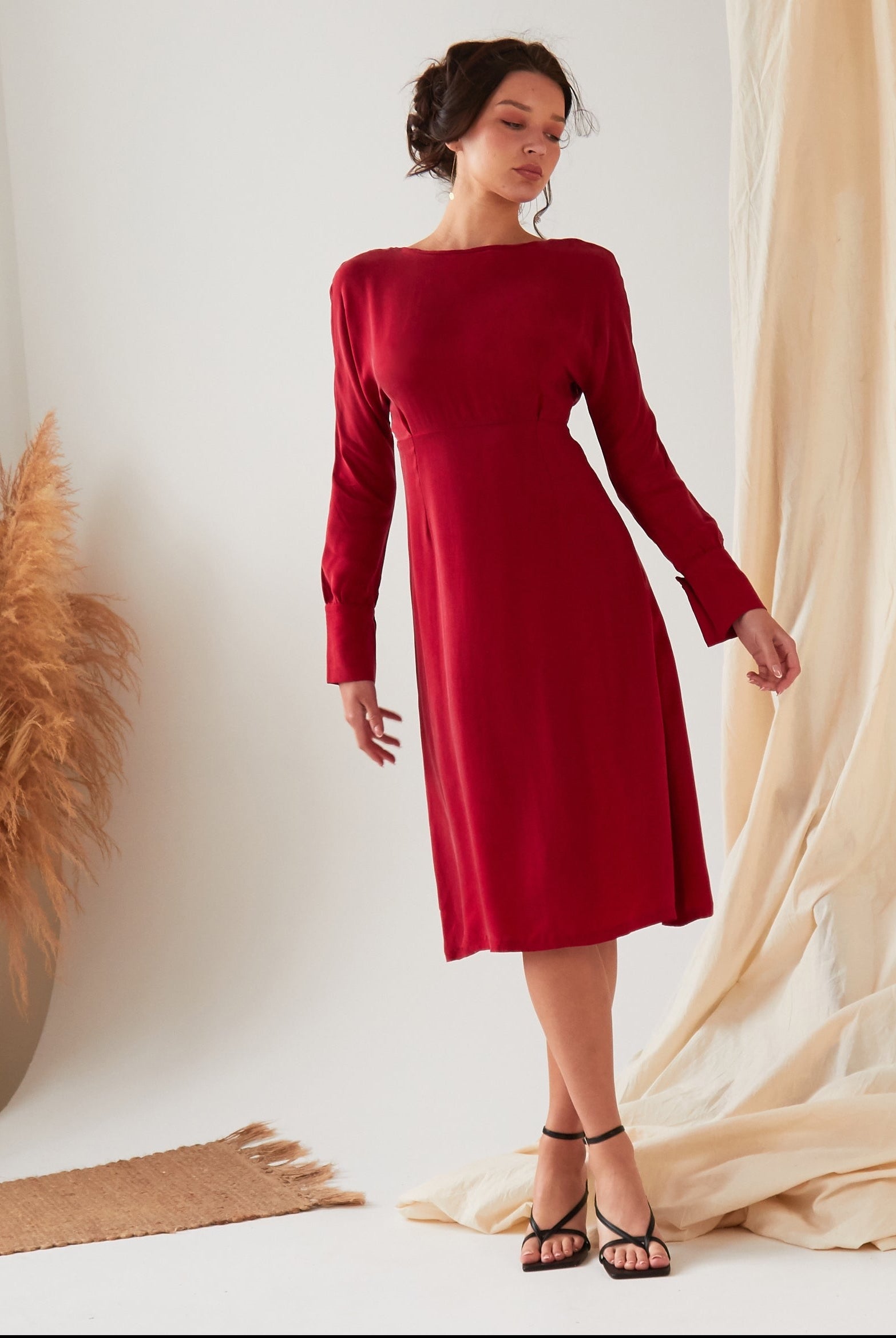 A woman stands indoors wearing the Sarvin Long Sleeve Red Midi Dress and black sandals. Beige fabric drapes and dried pampas grass decorate the background as she looks to the side with a relaxed posture.