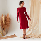 A woman stands indoors wearing the Sarvin Long Sleeve Red Midi Dress and black sandals. Beige fabric drapes and dried pampas grass decorate the background as she looks to the side with a relaxed posture.