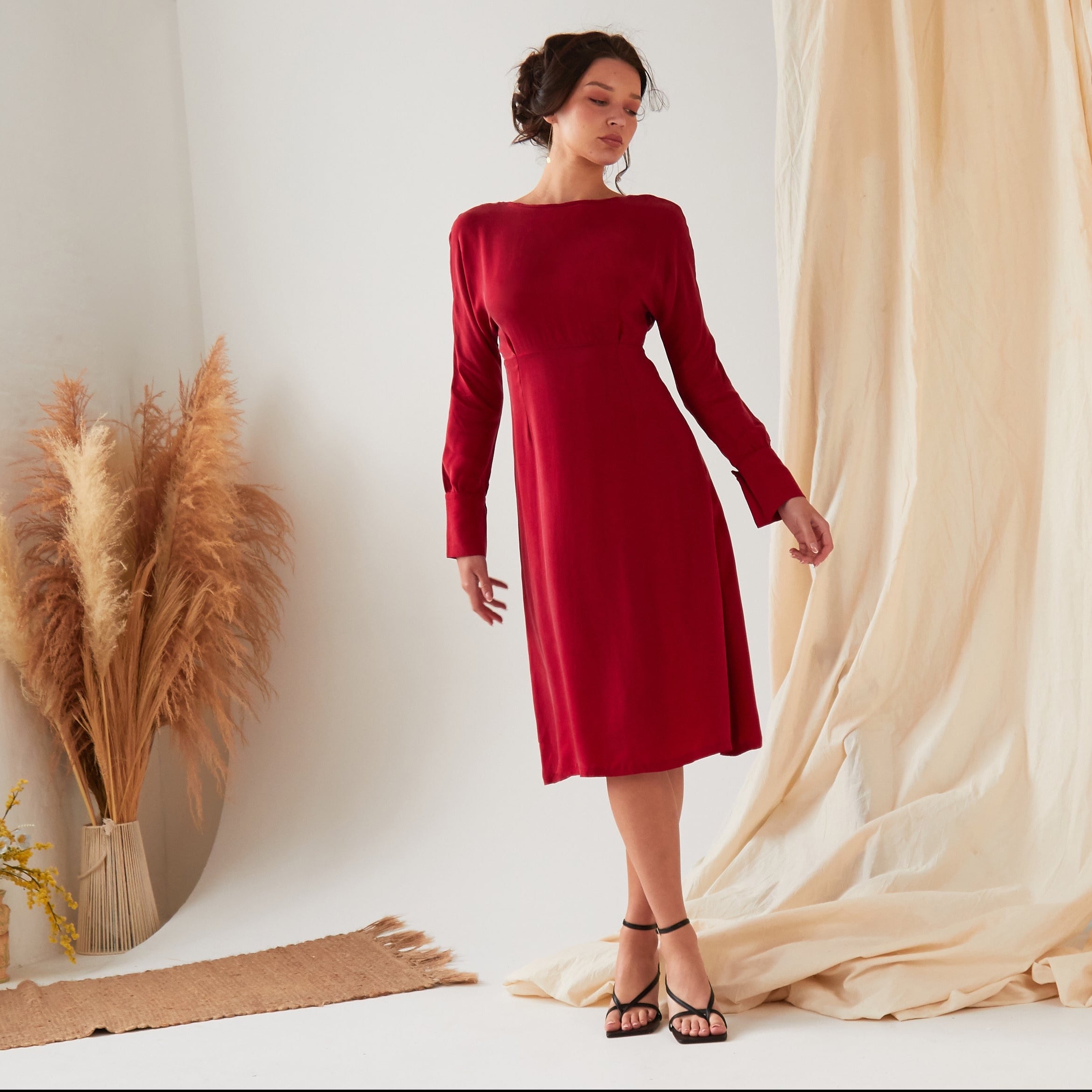 A woman stands indoors wearing the Sarvin Long Sleeve Red Midi Dress and black sandals. Beige fabric drapes and dried pampas grass decorate the background as she looks to the side with a relaxed posture.