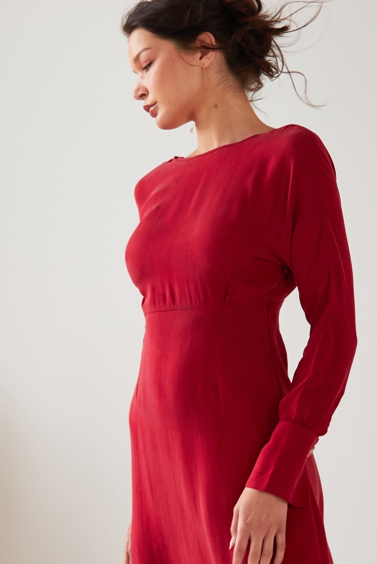A woman with dark hair tied up stands in profile wearing the Sarvin Long Sleeve Red Midi Dress. The backdrop is plain and light, with part of a plant on the left.