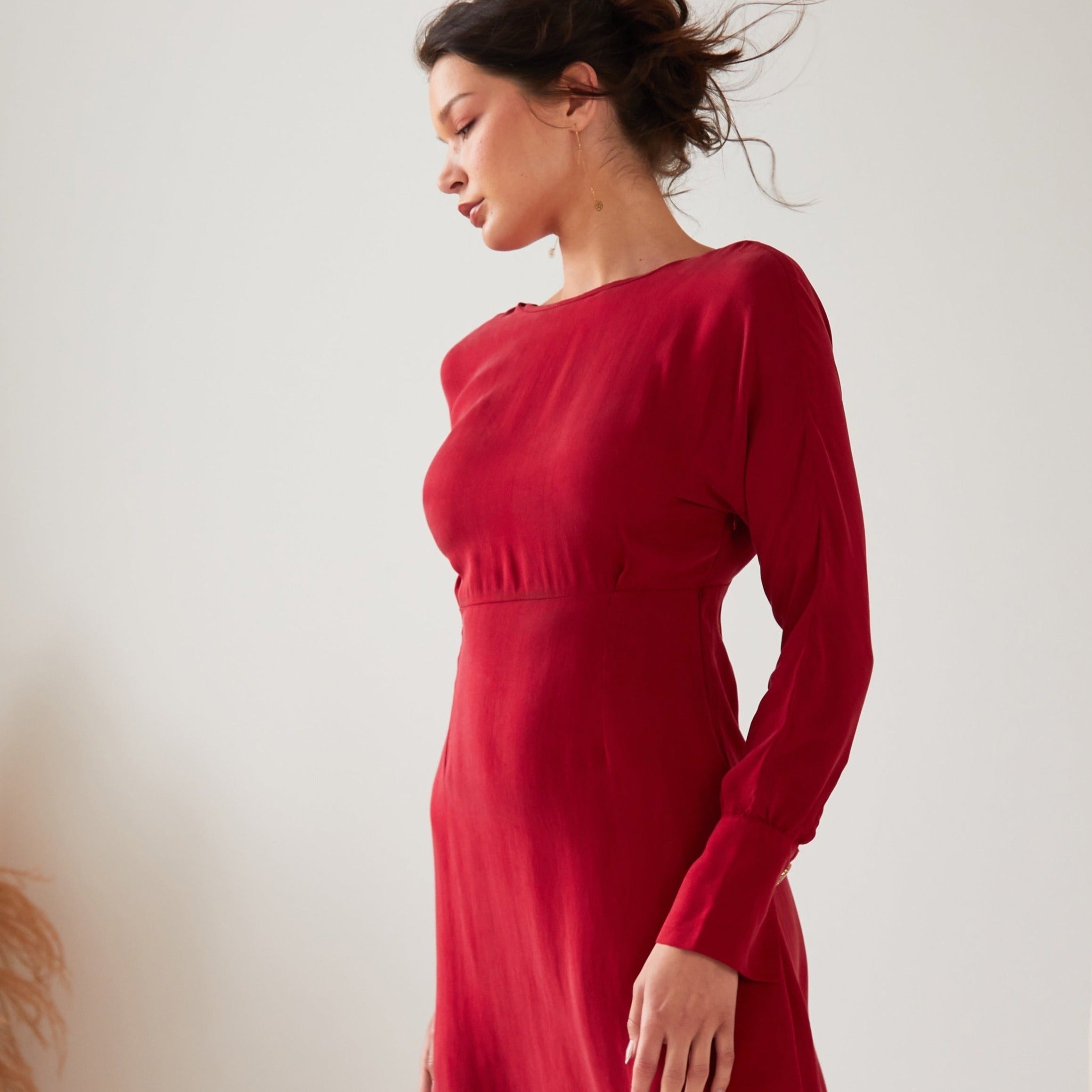 A woman with dark hair tied up stands in profile wearing the Sarvin Long Sleeve Red Midi Dress. The backdrop is plain and light, with part of a plant on the left.