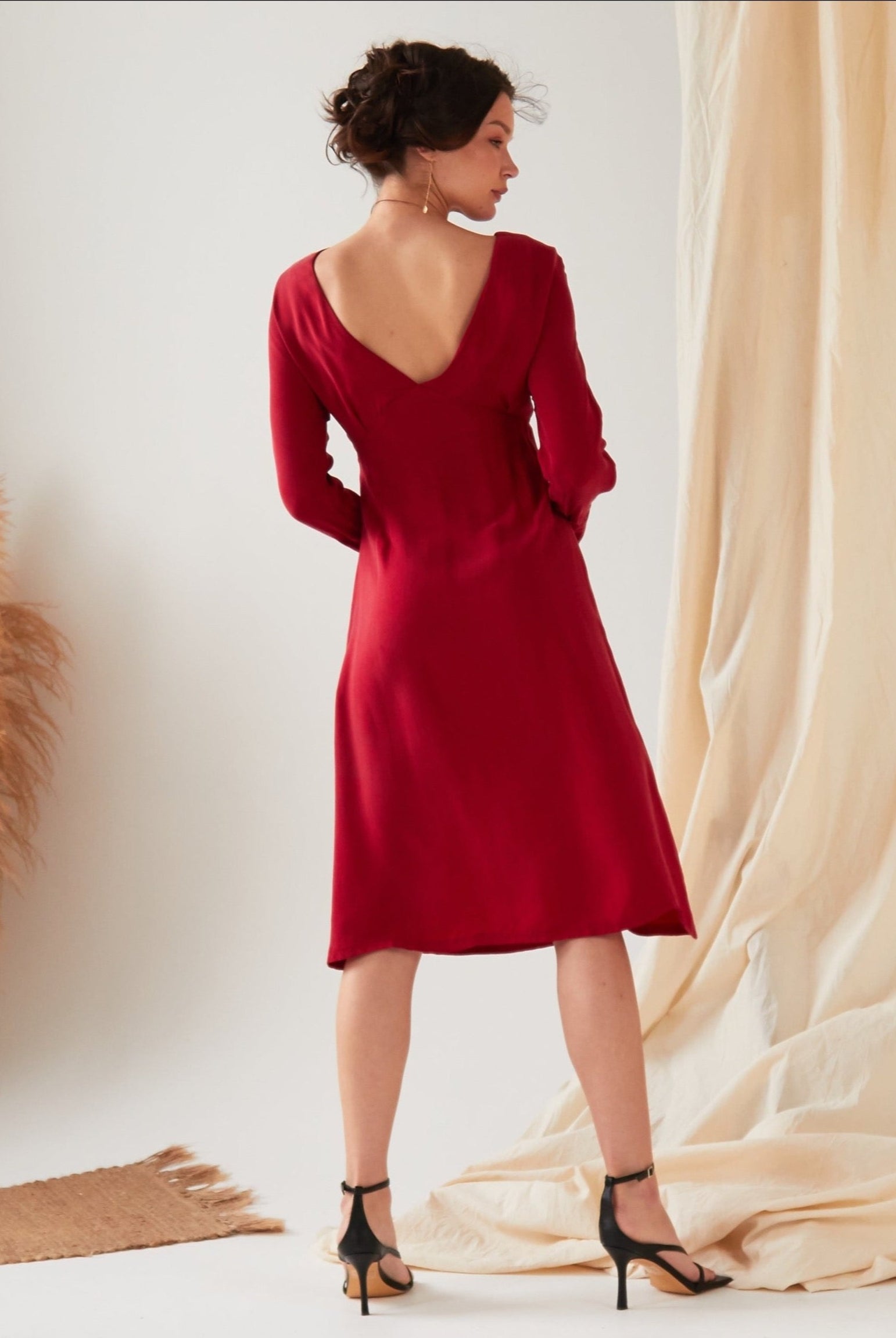 A woman stands indoors with her back to the camera, wearing the Sarvin Long Sleeve Red Midi Dress featuring a deep V-back. The scene includes a light floor, pampas grass, and beige fabric in the background.