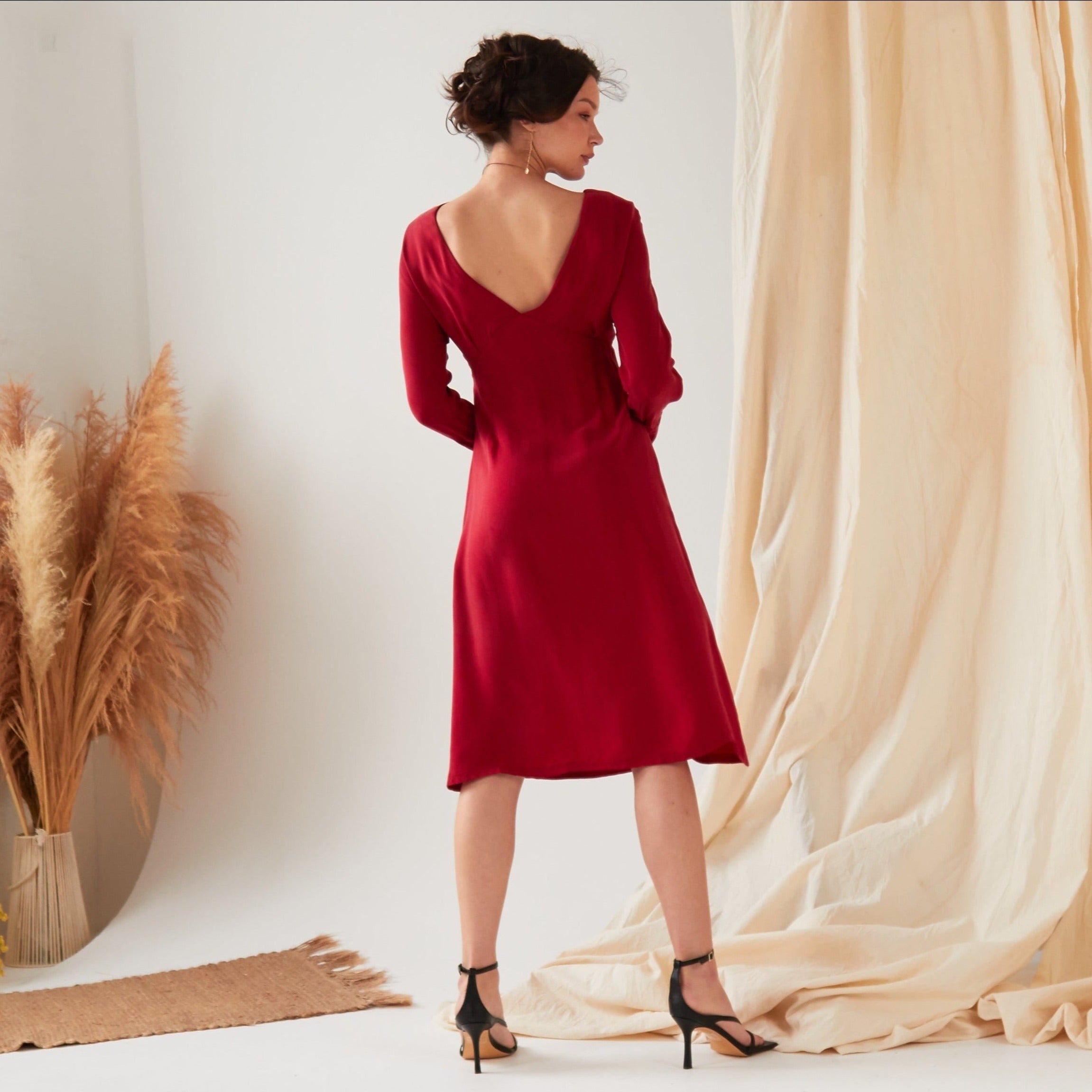 A woman stands indoors with her back to the camera, wearing the Sarvin Long Sleeve Red Midi Dress featuring a deep V-back. The scene includes a light floor, pampas grass, and beige fabric in the background.