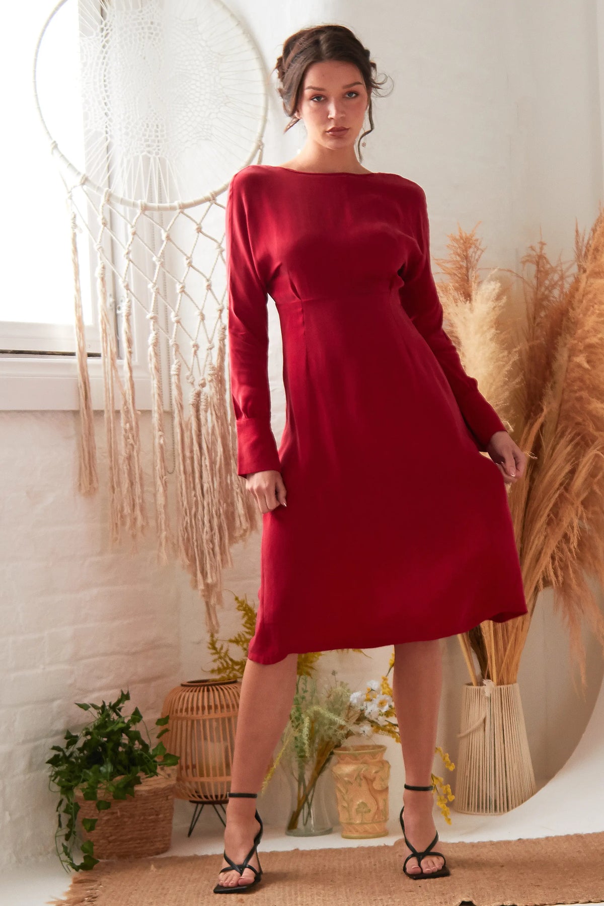A woman stands indoors in the Sarvin Long Sleeve Red Midi Dress and black strappy heels, with dried plants, a large dreamcatcher, and woven baskets against white brick walls and a window in the background.