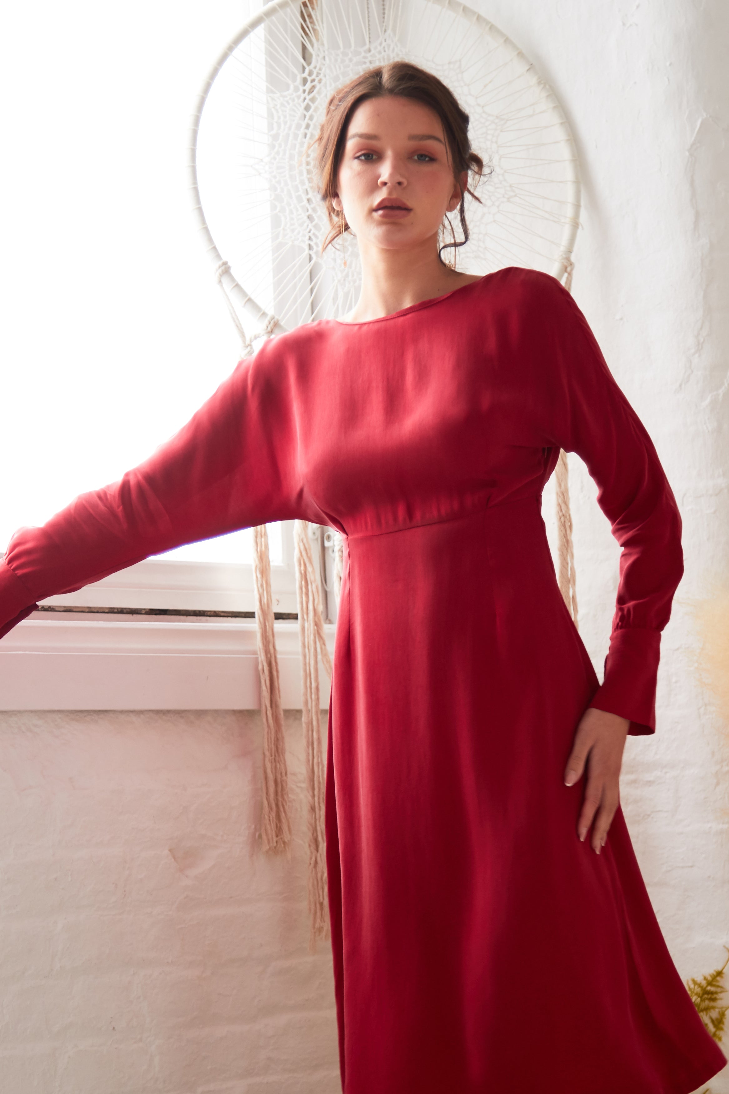 A person wears the Sarvin Long Sleeve Red Midi Dress indoors near a window with white walls, enhanced by a decorative white dreamcatcher hanging on the wall behind them.