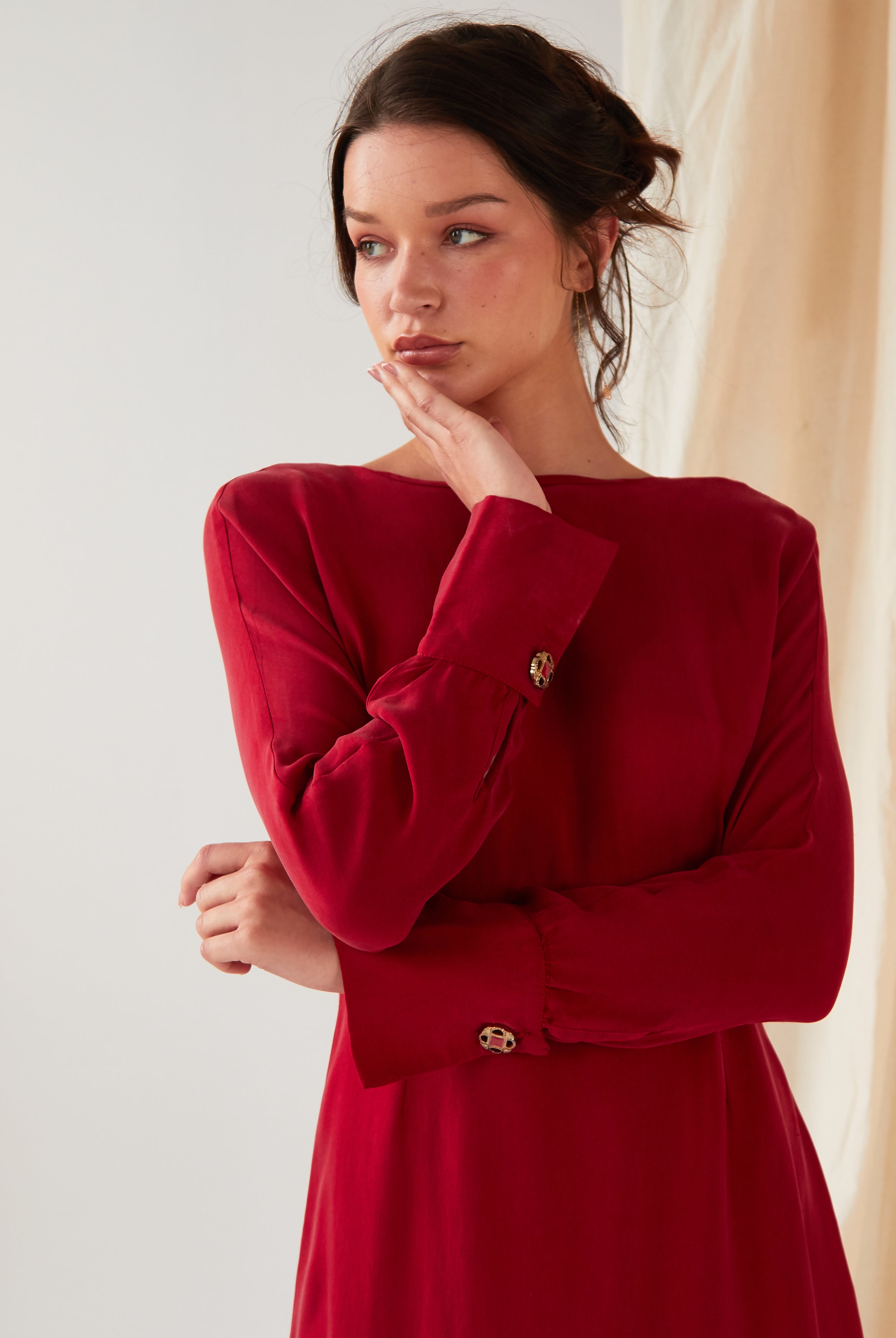 A woman wearing the Sarvin Long Sleeve Red Midi Dress stands against a light background, chin resting on her right hand and gazing left with a neutral expression. Her hair is loosely tied back.