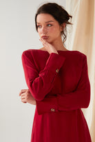 A woman wearing the Sarvin Long Sleeve Red Midi Dress stands against a light background, chin resting on her right hand and gazing left with a neutral expression. Her hair is loosely tied back.