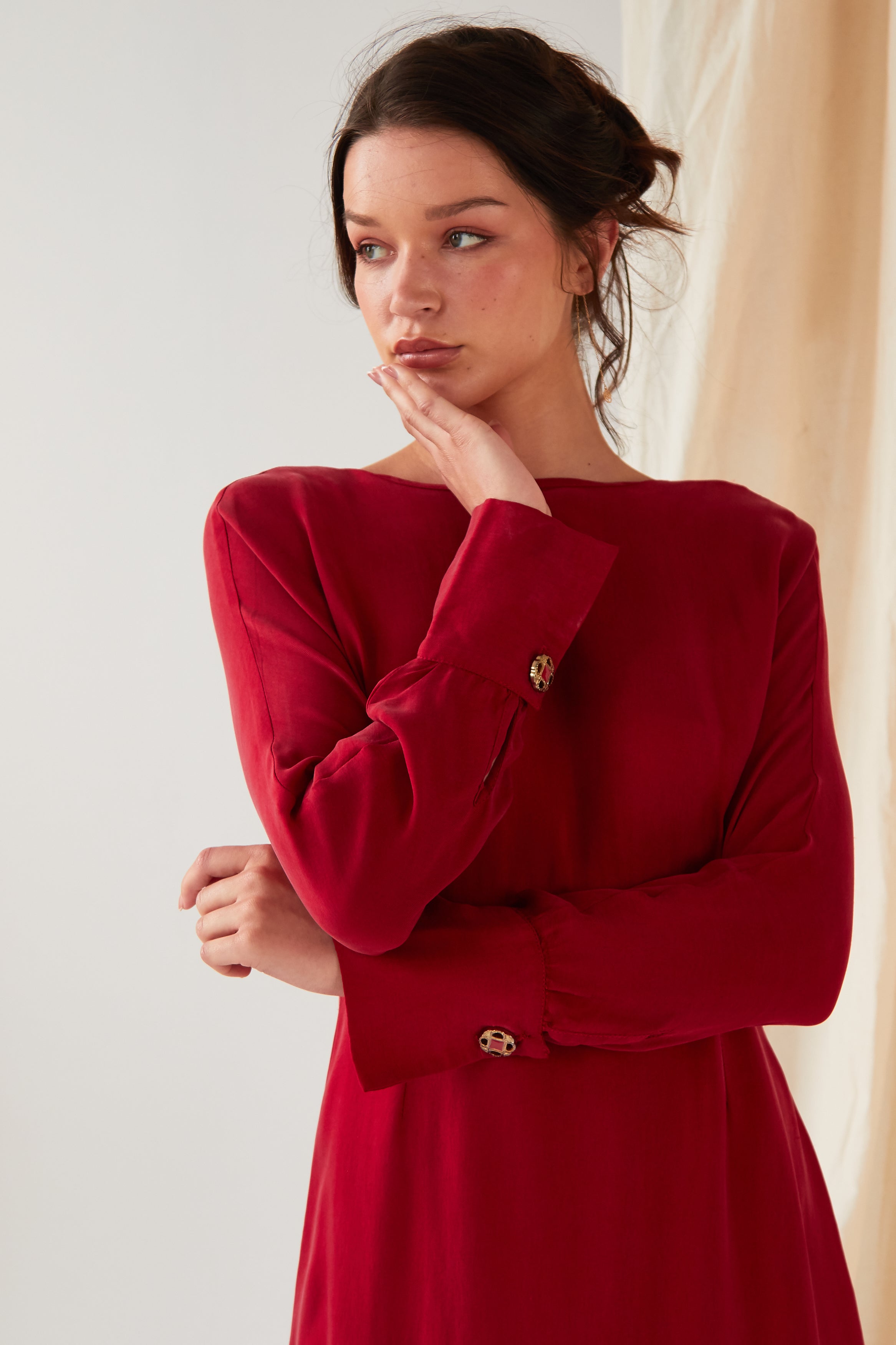 A woman wearing the Sarvin Long Sleeve Red Midi Dress stands against a light background, chin resting on her right hand and gazing left with a neutral expression. Her hair is loosely tied back.