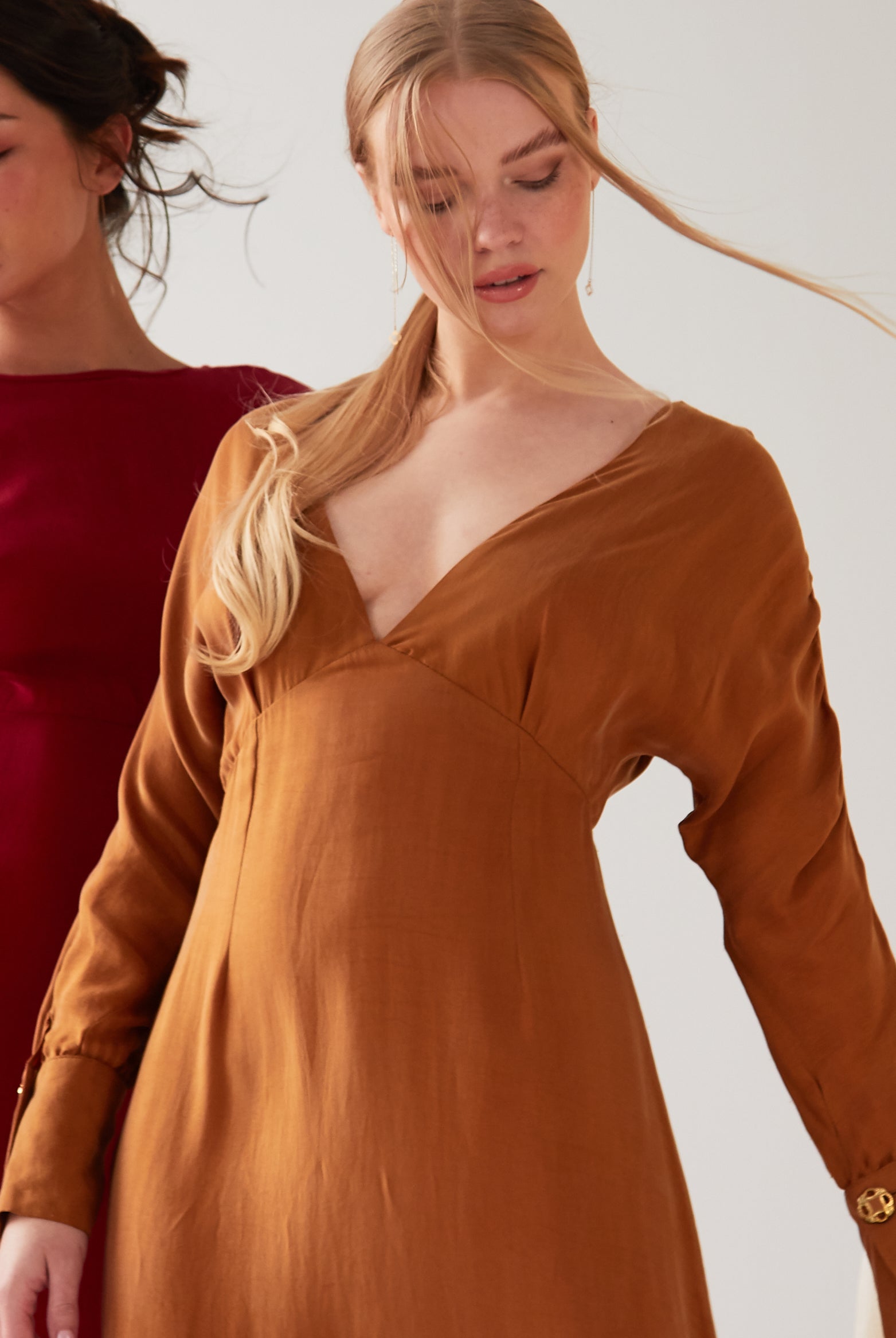Two women stand indoors wearing long-sleeved dresses: one in deep red Cupro, the other in the Sarvin Mustard Yellow Midi Dress. Both have light makeup and relaxed hair, posed in front of neutral-toned drapes.