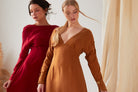 Two women stand indoors wearing long-sleeved dresses: one in deep red Cupro, the other in the Sarvin Mustard Yellow Midi Dress. Both have light makeup and relaxed hair, posed in front of neutral-toned drapes.