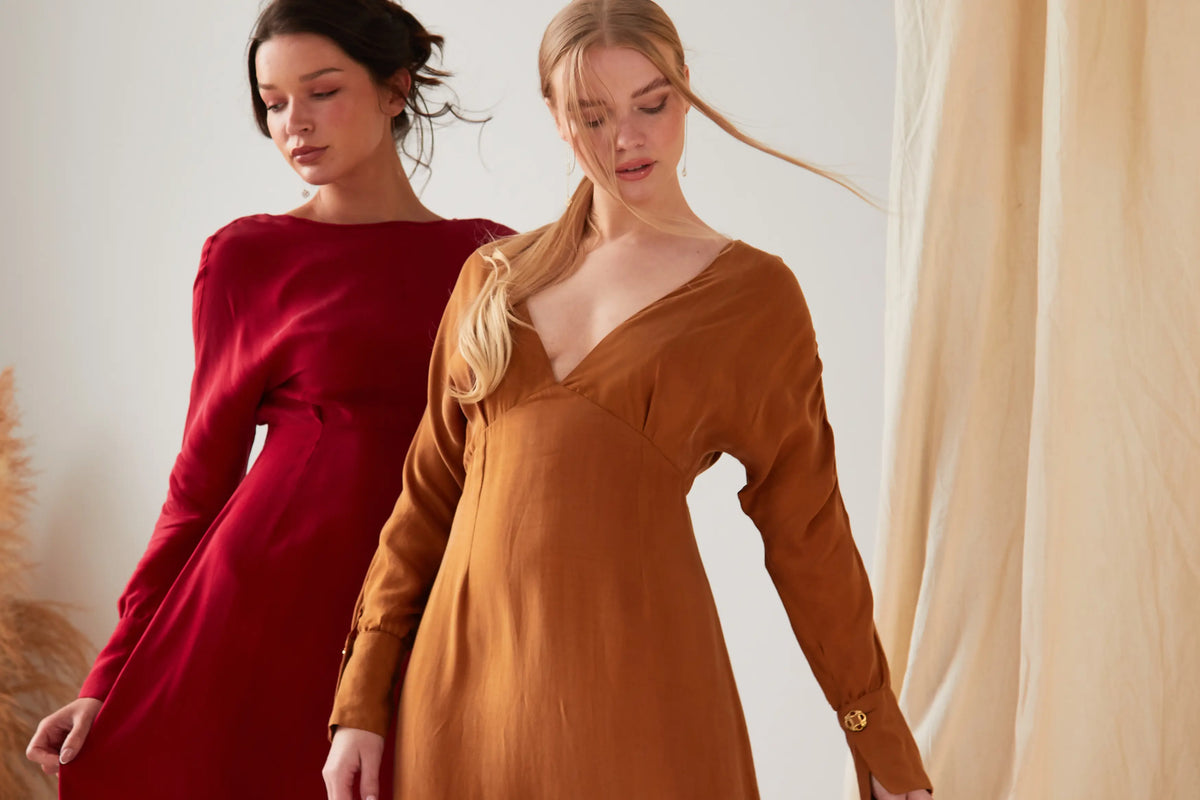 Two women stand indoors wearing long-sleeved dresses: one in deep red Cupro, the other in the Sarvin Mustard Yellow Midi Dress. Both have light makeup and relaxed hair, posed in front of neutral-toned drapes.