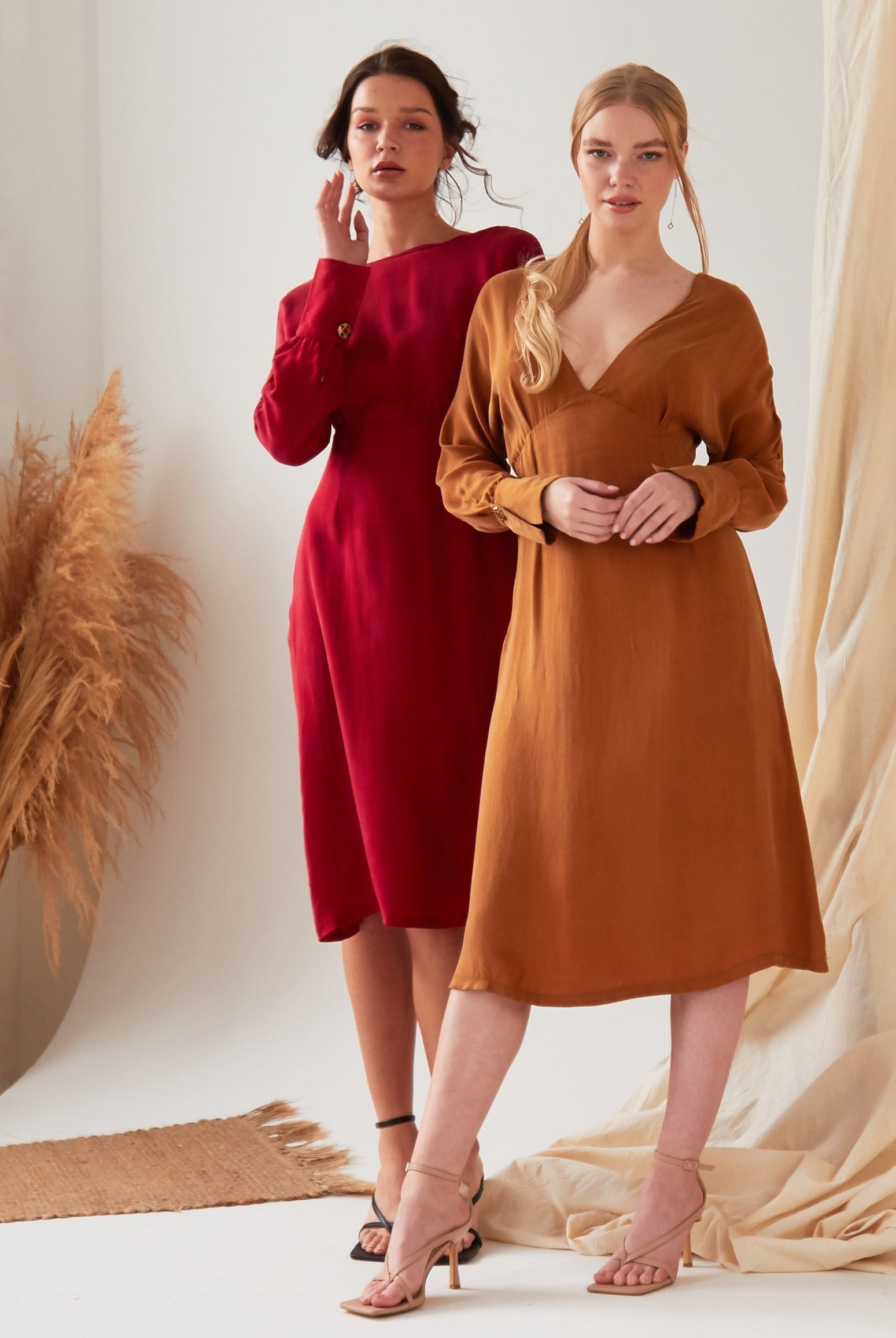 Two women pose confidently in a minimal studio with pampas grass and draped fabric. One wears the Sarvin Mustard Yellow Midi Dress, while the other sports a brown long-sleeve v-neck style; both pair their looks with heels.
