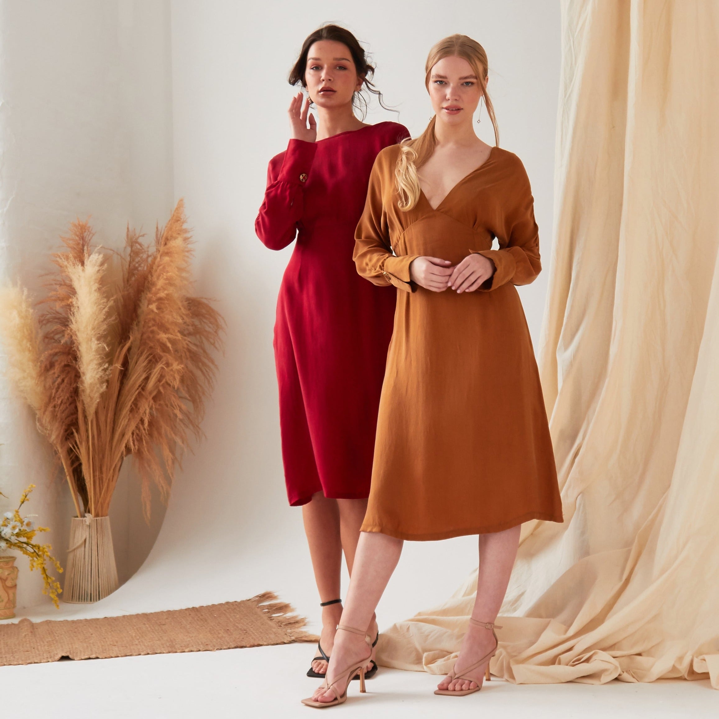Two women pose confidently in a minimal studio with pampas grass and draped fabric. One wears the Sarvin Mustard Yellow Midi Dress, while the other sports a brown long-sleeve v-neck style; both pair their looks with heels.