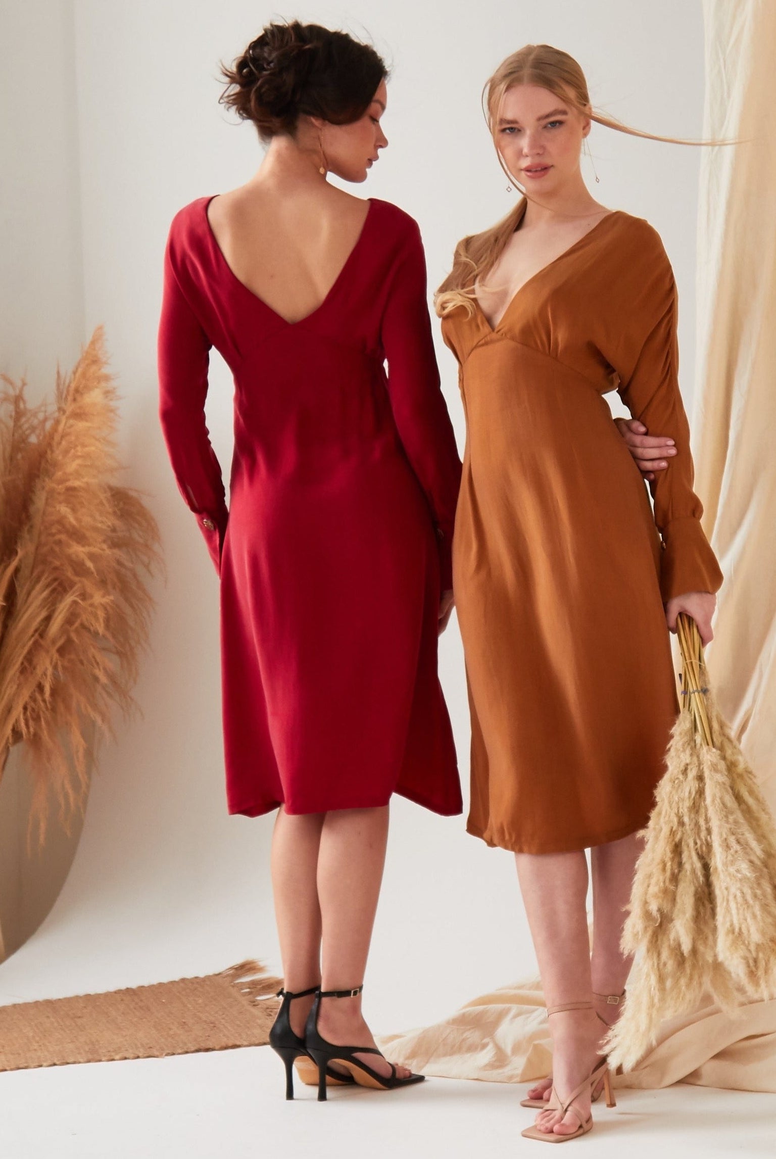 Two women indoors stand amid neutral drapes. One, facing away, wears the Sarvin Long Sleeve Red Midi Dress with black heels; the other faces forward in a brown eco-friendly Cupro dress and holds pampas grass.