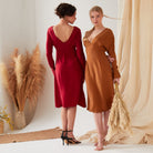 Two women indoors stand amid neutral drapes. One, facing away, wears the Sarvin Long Sleeve Red Midi Dress with black heels; the other faces forward in a brown eco-friendly Cupro dress and holds pampas grass.