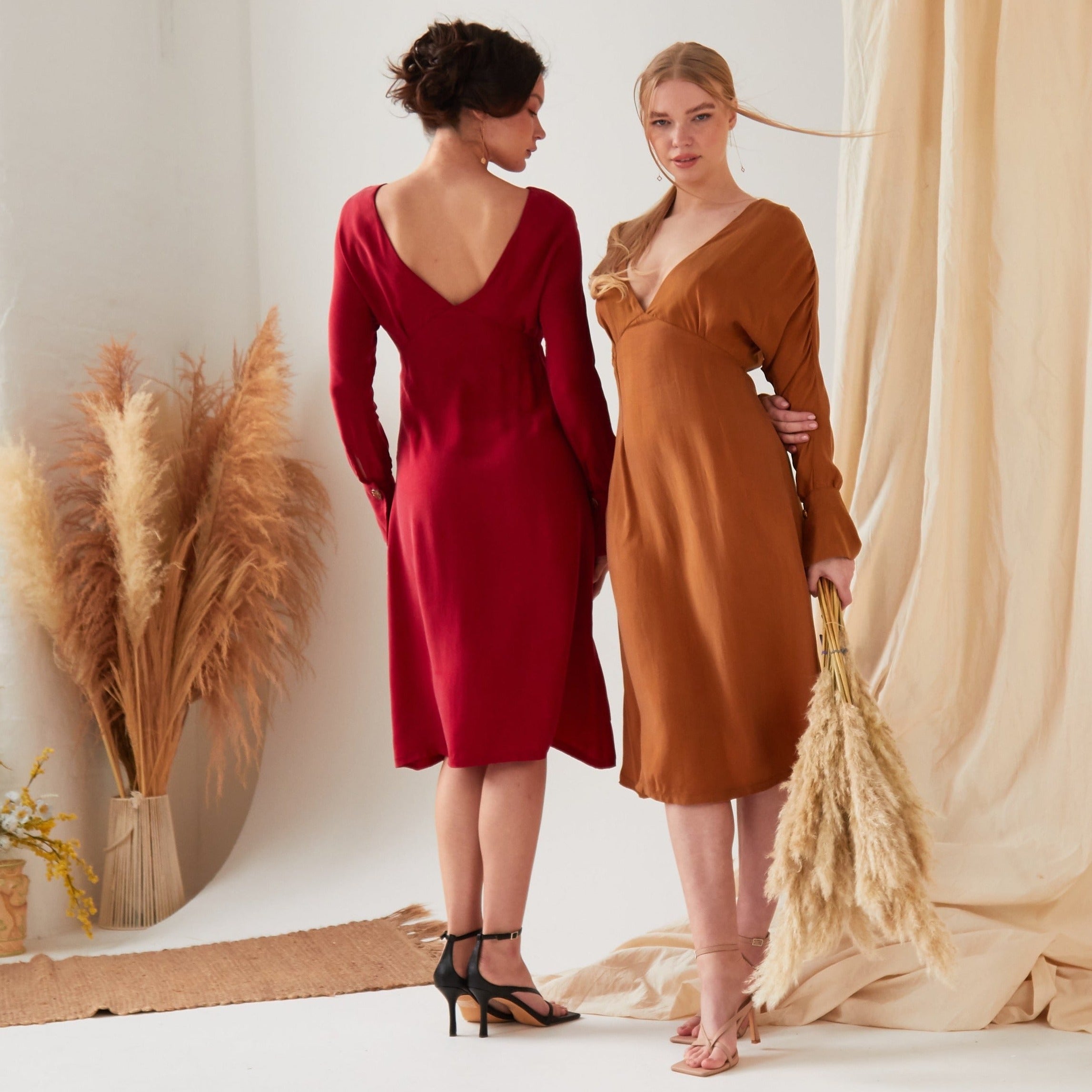 Two women indoors stand amid neutral drapes. One, facing away, wears the Sarvin Long Sleeve Red Midi Dress with black heels; the other faces forward in a brown eco-friendly Cupro dress and holds pampas grass.
