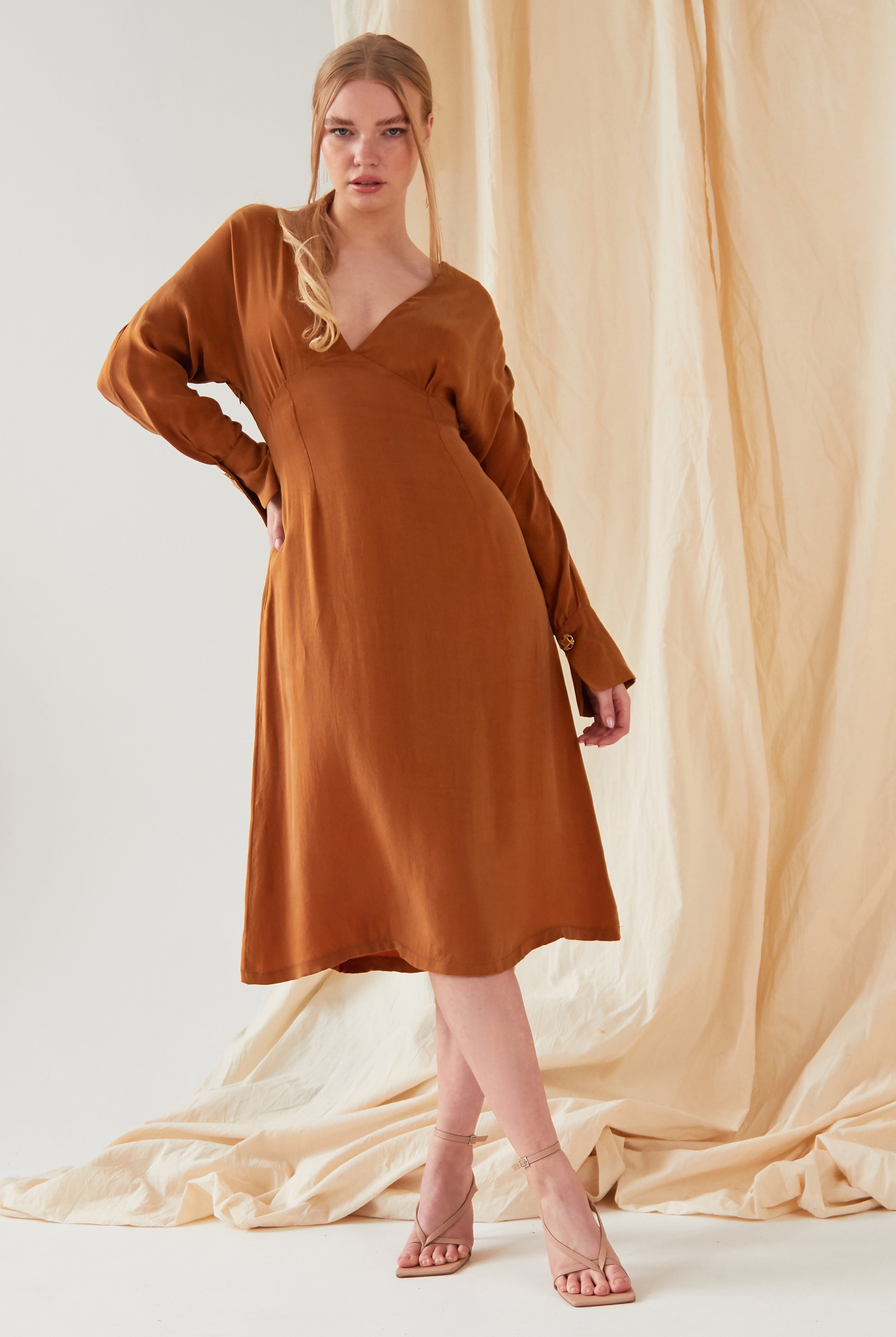 A woman stands in front of beige fabric wearing the Sarvin Mustard Yellow Midi Dress with nude strappy heels. Her blonde hair is styled in a loose ponytail as she looks directly at the camera.