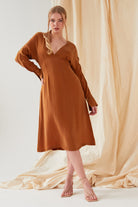 A woman stands in front of beige fabric wearing the Sarvin Mustard Yellow Midi Dress with nude strappy heels. Her blonde hair is styled in a loose ponytail as she looks directly at the camera.