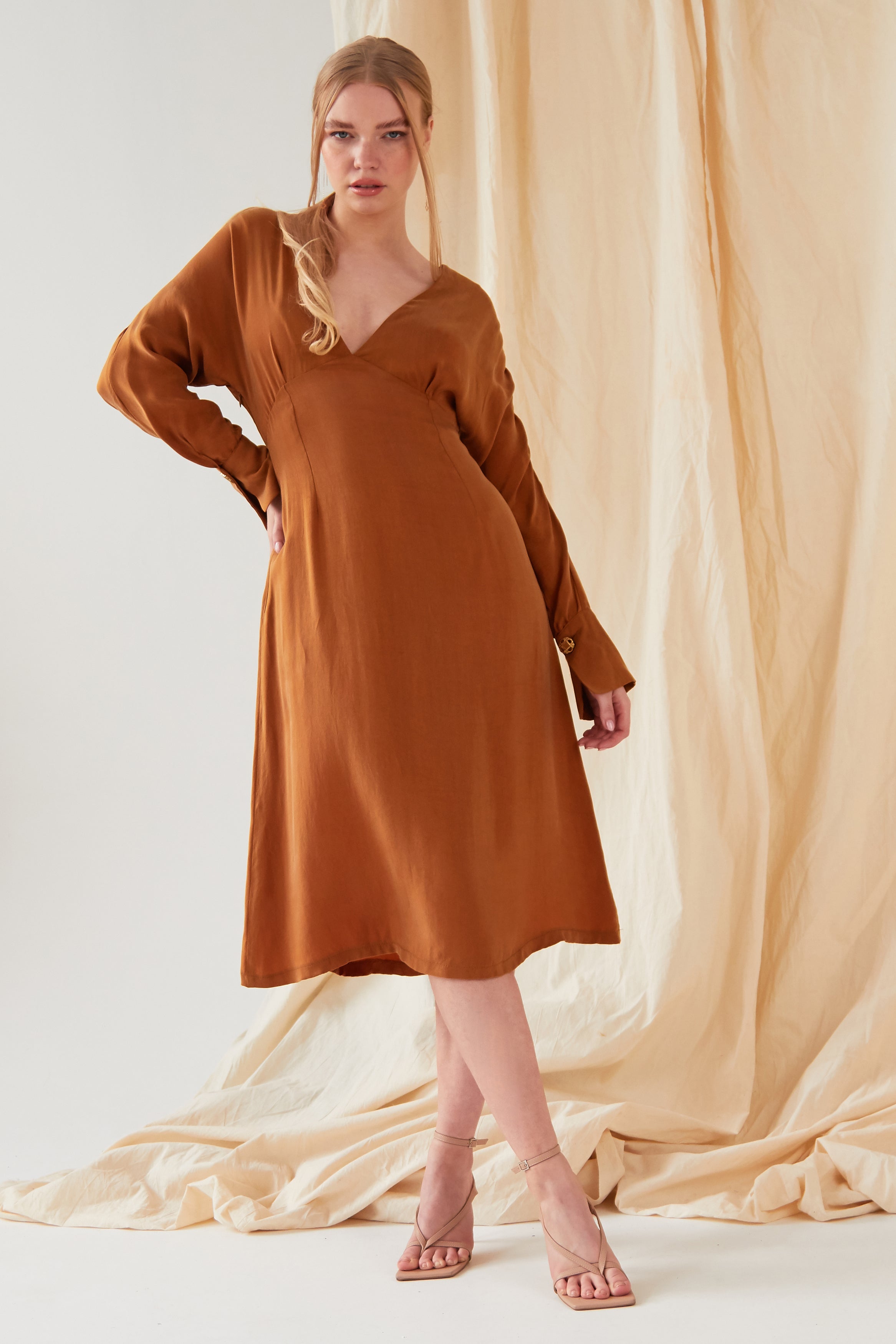 A woman stands in front of beige fabric wearing the Sarvin Mustard Yellow Midi Dress with nude strappy heels. Her blonde hair is styled in a loose ponytail as she looks directly at the camera.