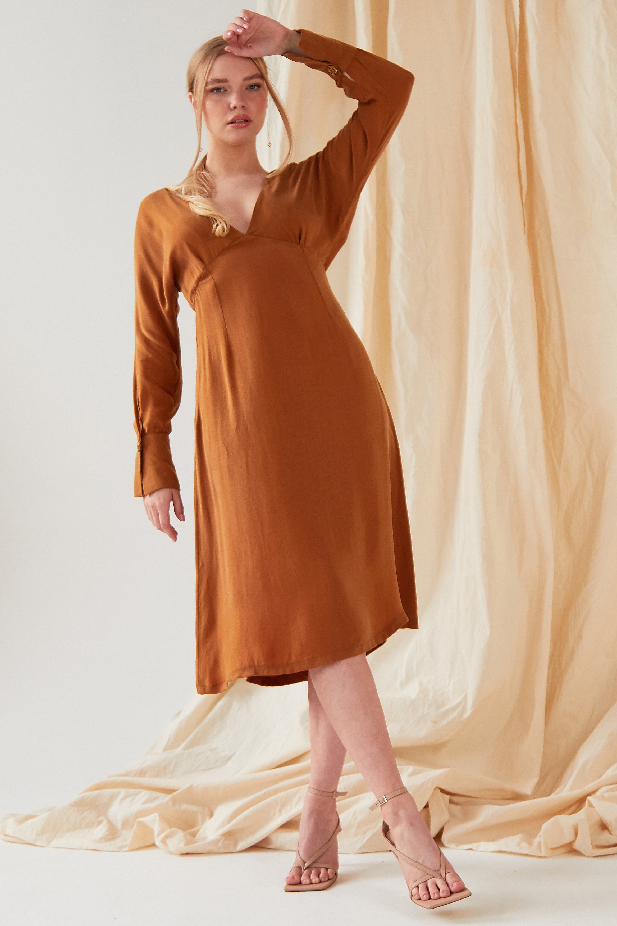 A person with light hair stands in front of a beige fabric backdrop, wearing the Sarvin Mustard Yellow Midi Dress and strappy sandals, with one arm raised and resting on their head.