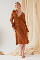 A person wearing the Sarvin Mustard Yellow Midi Dress with long sleeves and a V-neckline, styled with strappy sandals, stands before draped beige fabric.