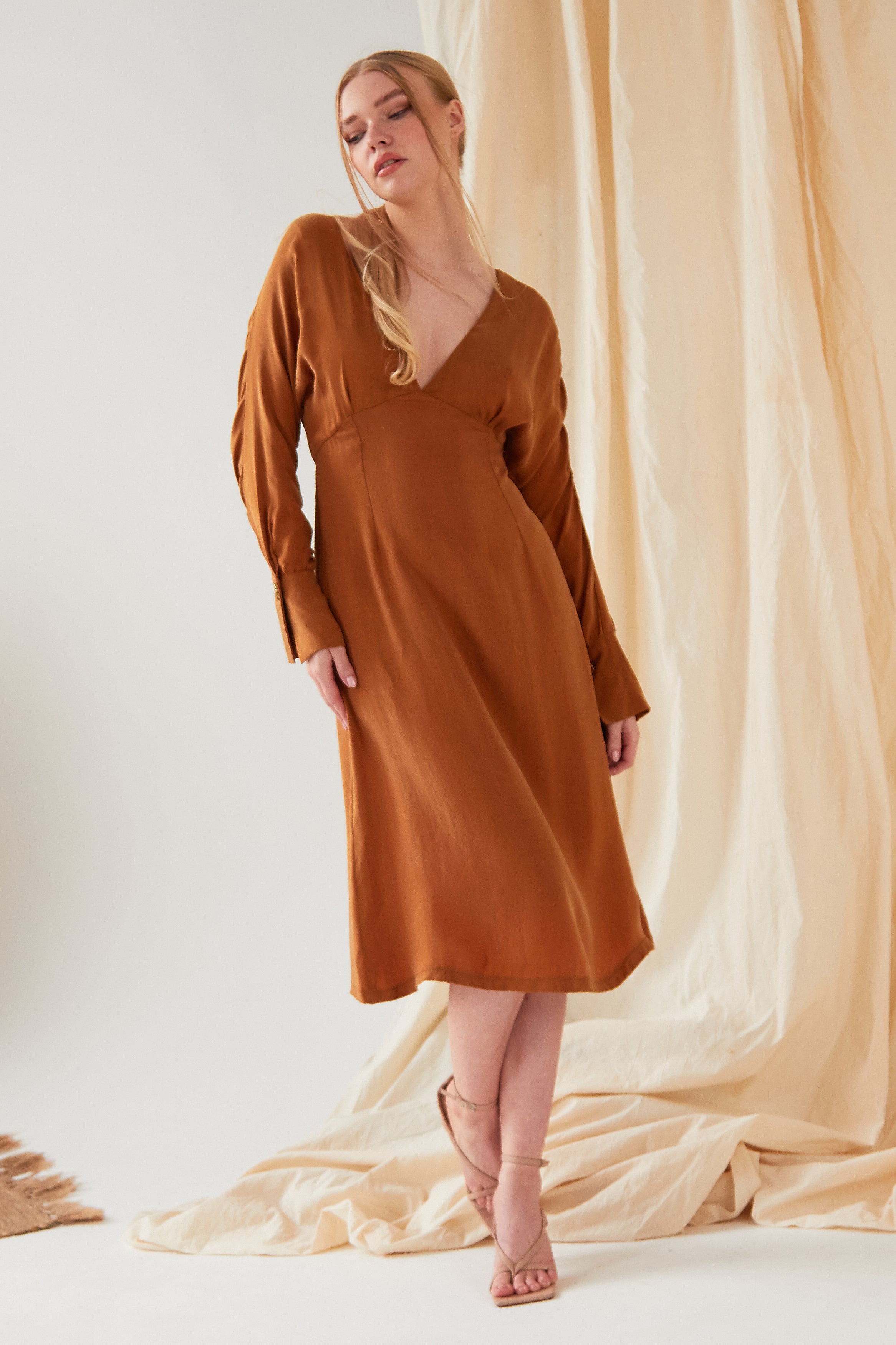 A person wearing the Sarvin Mustard Yellow Midi Dress with long sleeves and a V-neckline, styled with strappy sandals, stands before draped beige fabric.
