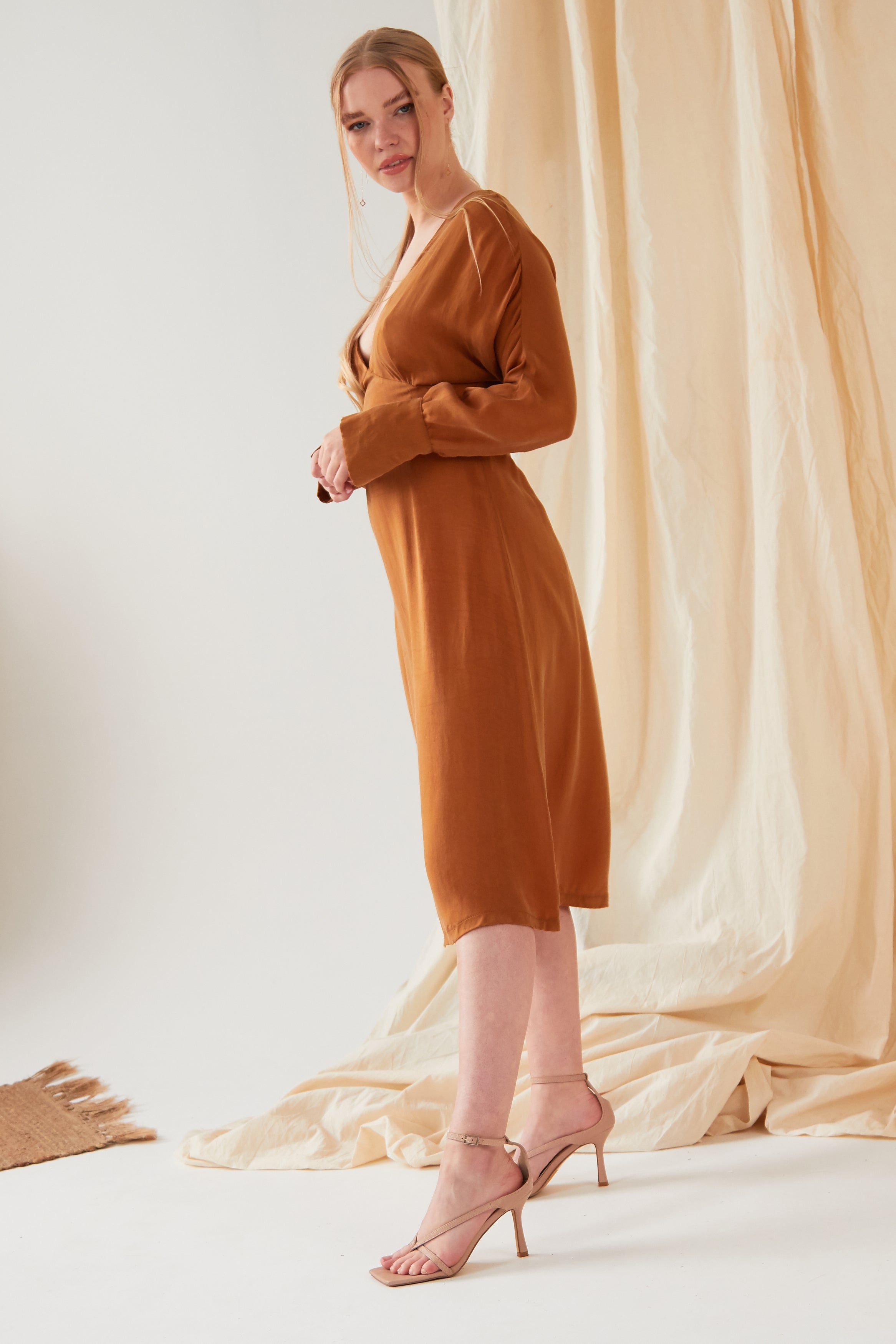 A woman stands sideways indoors, wearing the Sarvin Mustard Yellow Midi Dress with long sleeves and knee-length hem, paired with beige high-heeled sandals. A beige draped fabric backdrop and a woven rug are partially visible in the scene.