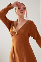 A woman wears the Sarvin Mustard Yellow Midi Dress with long sleeves, standing indoors against a plain light background, one arm raised and her blonde hair styled in a loose ponytail.