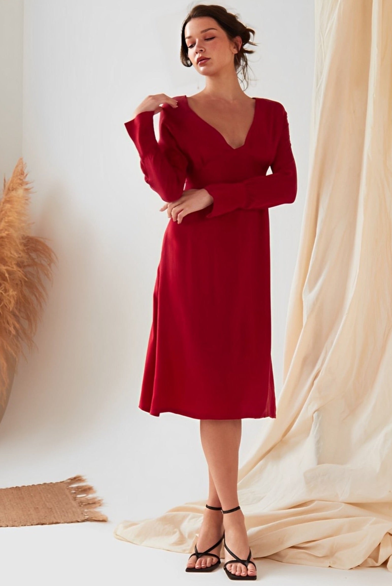 A woman wearing the Sarvin Long Sleeve Red Midi Dress stands indoors near beige curtains and a vase of pampas grass.