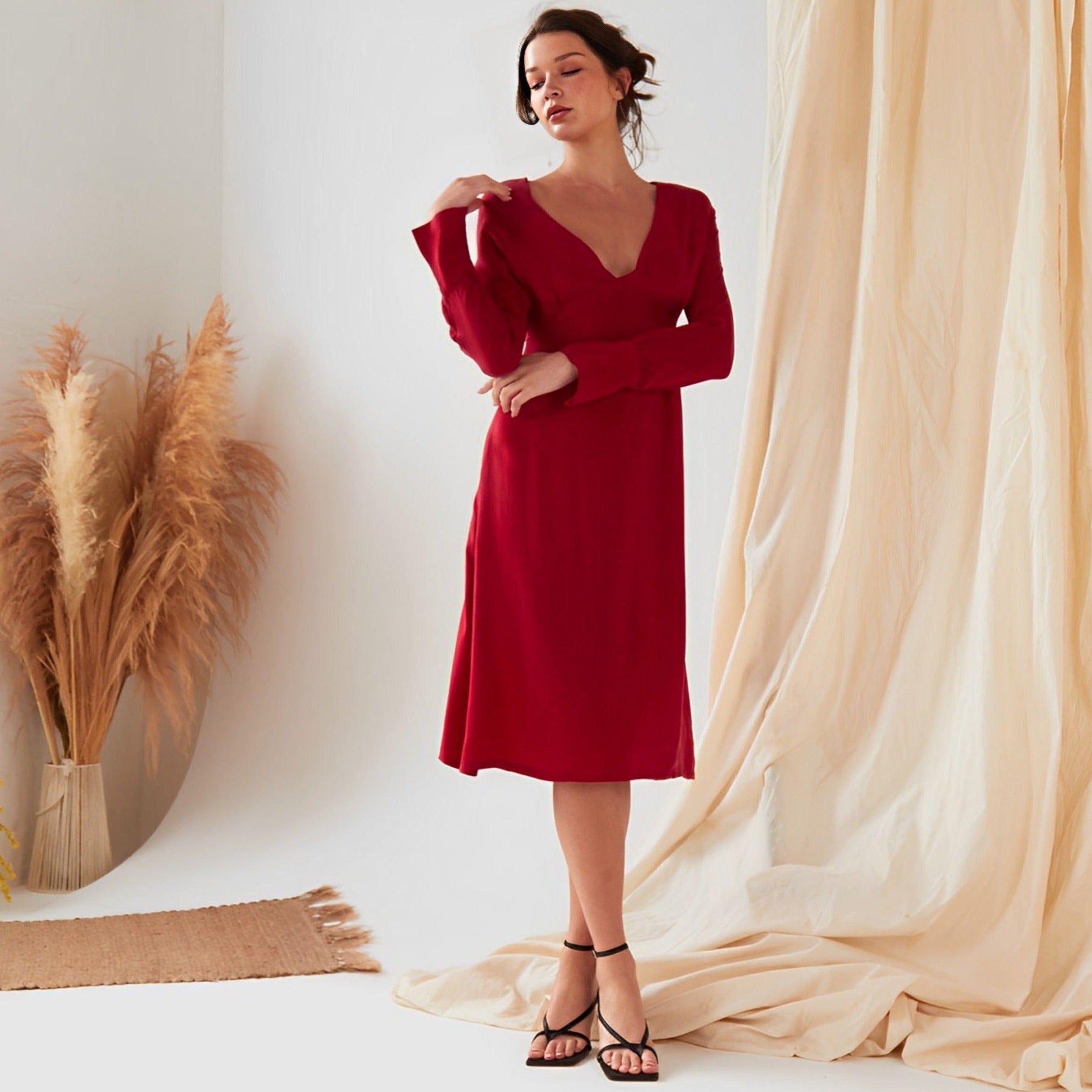A woman wearing the Sarvin Long Sleeve Red Midi Dress stands indoors near beige curtains and a vase of pampas grass.