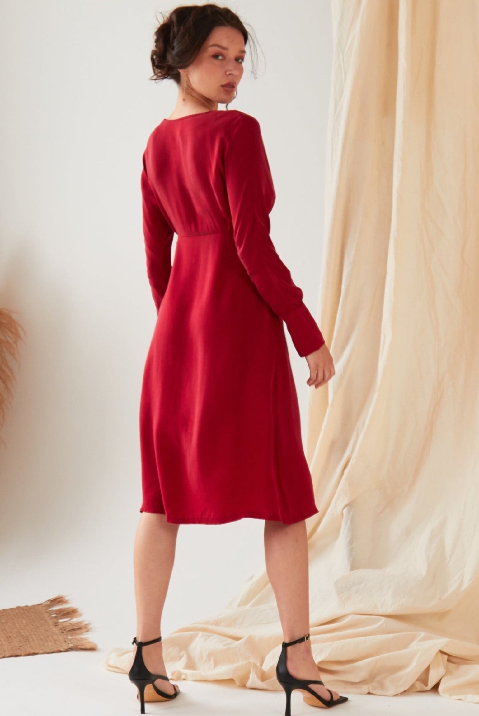 A woman with dark hair in an updo wears the Sarvin Long Sleeve Red Midi Dress and black heels, standing on a light floor before beige curtains, with dried pampas grass beside her.