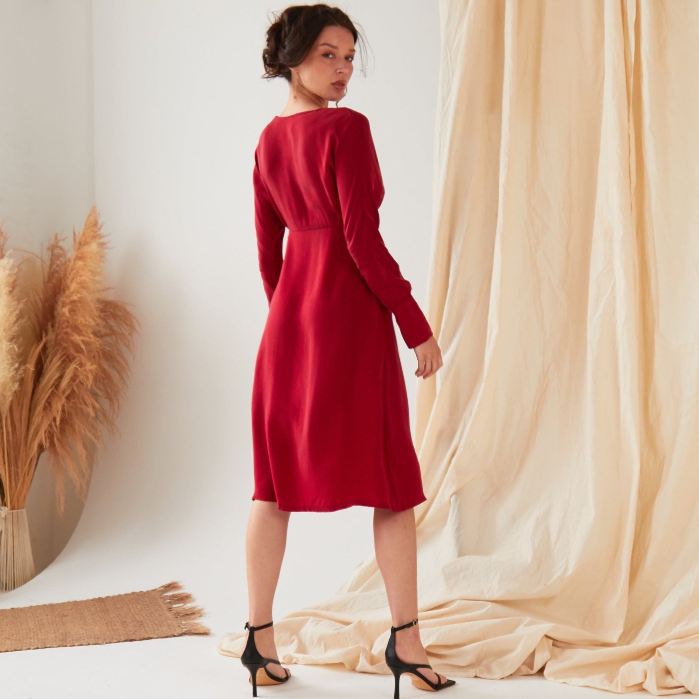A woman with dark hair in an updo wears the Sarvin Long Sleeve Red Midi Dress and black heels, standing on a light floor before beige curtains, with dried pampas grass beside her.