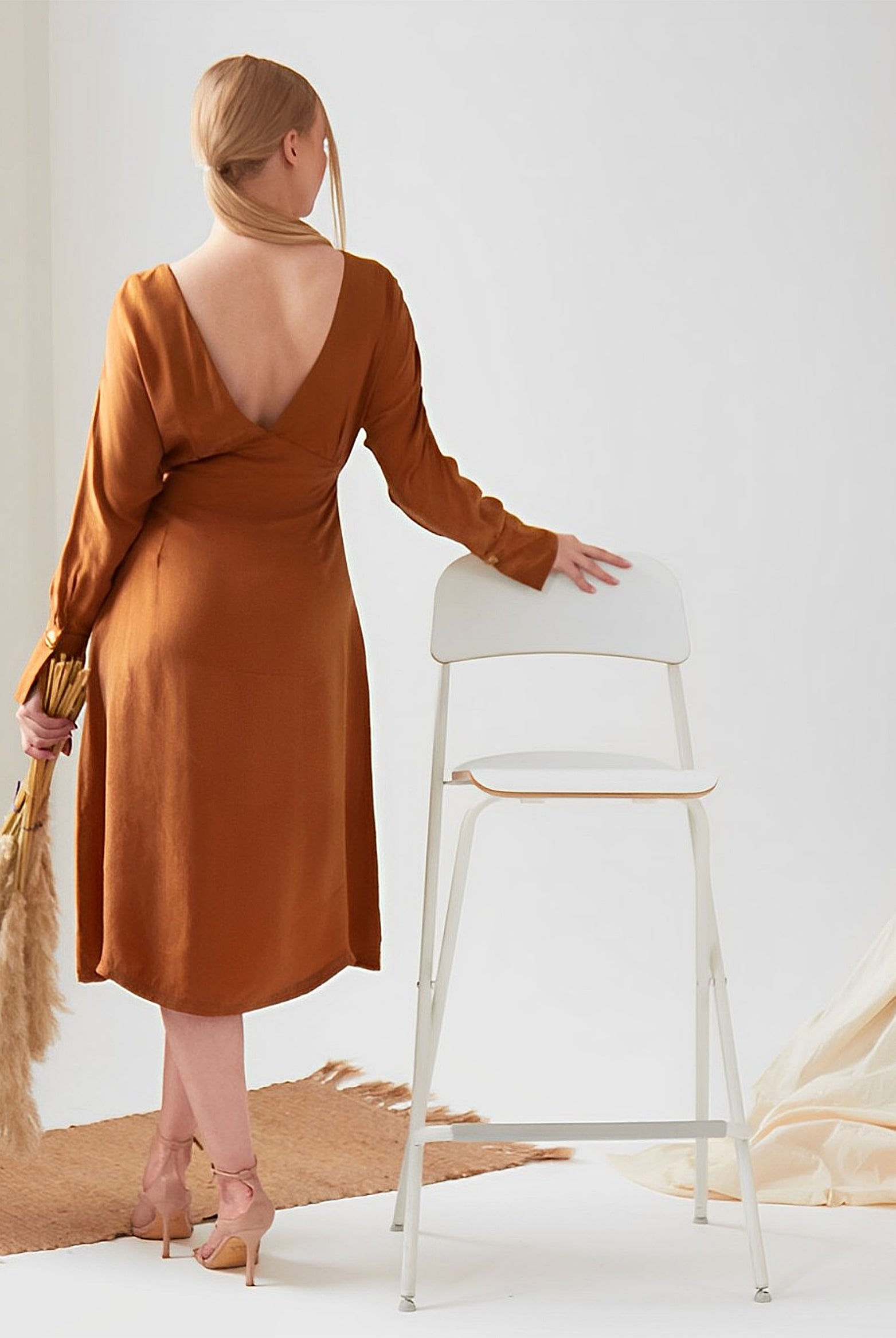 A woman in a Sarvin Mustard Yellow Midi Dress stands with her back to the camera, holding pampas grass and resting her hand on a white chair in a softly decorated, neutral-toned room.